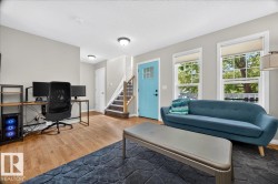 Bright, open living space featuring hardwood flooring, abundant natural light from multiple large windows, a blue upholstered sofa, and a dedicated workspace with built-in desk and shelving - 