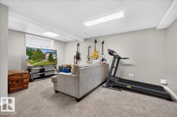 Versatile recreation room featuring neutral carpeting, recessed and linear ceiling lighting, built-in wall-mounted guitar display with multiple hanging mounts, gray upholstered seating, wood-finish media console, wall-mounted television, and a treadmill - 
