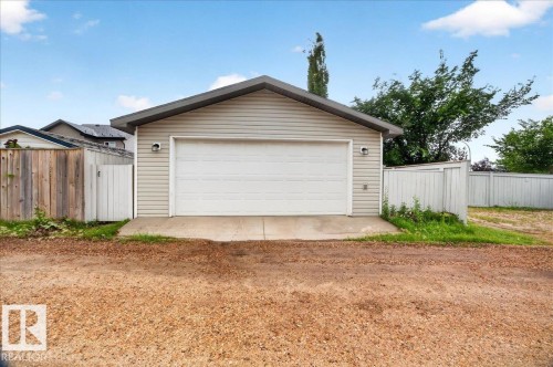 Spacious two-car garage featuring a white paneled overhead door and concrete apron, complemented by vinyl siding exterior and mature landscaping with privacy fencing on either side - 887 Oakland Boulevard, Devon, AB - Outdoor
