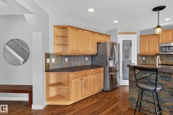 Open-concept kitchen featuring wood-finish flooring, light wood cabinetry, stone-look tile backsplash, stainless steel appliances, and a breakfast bar with stacked stone facade - 