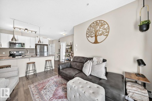 Open-concept living area featuring a modern kitchen with stainless steel appliances, white cabinetry, and a breakfast bar with seating, complemented by wood-finish flooring throughout - #238 1008 Rosenthal Bv Nw, Edmonton, AB - Indoor