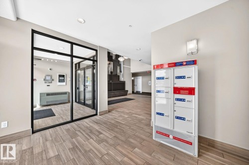 Modern commercial or professional space featuring a wood-finish herringbone floor, frameless glass entry doors with black aluminum framing, recessed ceiling lighting, and a wall-mounted organizational system with red and blue storage components - #238 1008 Rosenthal Bv Nw, Edmonton, AB - Indoor Photo Showing Other Room