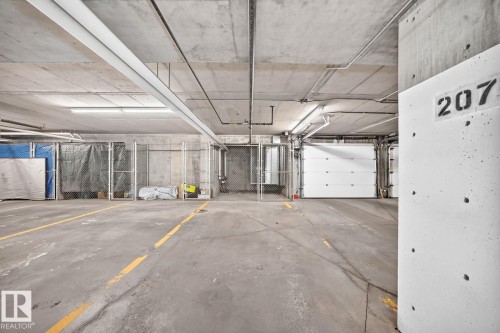 Concrete flooring with yellow directional markings, exposed concrete ceiling with utility conduits, fluorescent lighting, and a white panel garage door - #238 1008 Rosenthal Bv Nw, Edmonton, AB - Indoor Photo Showing Garage