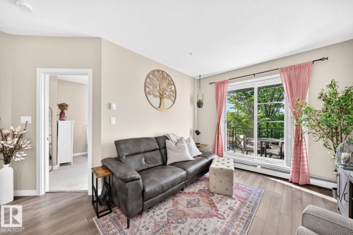 Bright living room showcasing hardwood flooring, a charcoal upholstered sofa, and expansive sliding glass doors opening to a private outdoor space - #238 1008 Rosenthal Bv Nw, Edmonton, AB - Indoor Photo Showing Living Room