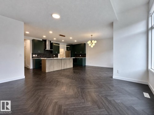 Spacious interior featuring dark wood-finish flooring in a herringbone pattern, an open-concept kitchen with dark cabinetry, a central island with a light-toned countertop, recessed ceiling lighting, and a modern globe chandelier - 5478 Kootook Road Sw, Edmonton, AB - Indoor