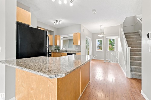 Kitchen island with granite countertop, wood-finish cabinetry, stainless steel appliances, and wood-finish flooring extending into the dining area with a stairway featuring carpeted treads and white railings - #144 104 West Haven Dr, Leduc, AB - Indoor Photo Showing Kitchen With Upgraded Kitchen