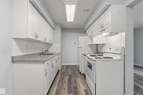 Galley-style kitchen featuring white cabinetry with chrome hardware, laminate countertops, subway tile backsplash, electric range with cooktop, refrigerator, and wood-finish flooring throughout - #9 14230 80 St Nw, Edmonton, AB - Indoor Photo Showing Kitchen