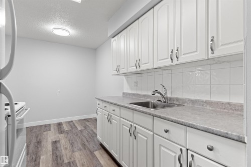 Wood-finish flooring with white baseboards, white shaker-style cabinetry, square tile backsplash, and a stainless steel sink with a gooseneck faucet - #9 14230 80 St Nw, Edmonton, AB - Indoor Photo Showing Kitchen