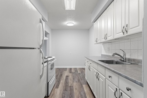Galley kitchen featuring white cabinetry with hardware accents, stainless steel appliances including a gas range and refrigerator, gray countertops, subway tile backsplash, and wood-finish flooring throughout - #9 14230 80 St Nw, Edmonton, AB - Indoor Photo Showing Kitchen