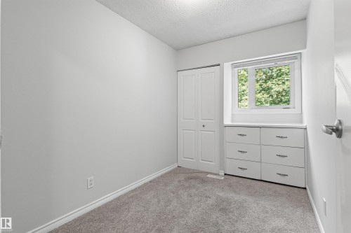 Bright secondary bedroom featuring neutral wall finishes, plush carpet flooring, a double closet with white panel doors, and a large window with natural light and exterior greenery views - #9 14230 80 St Nw, Edmonton, AB - Indoor