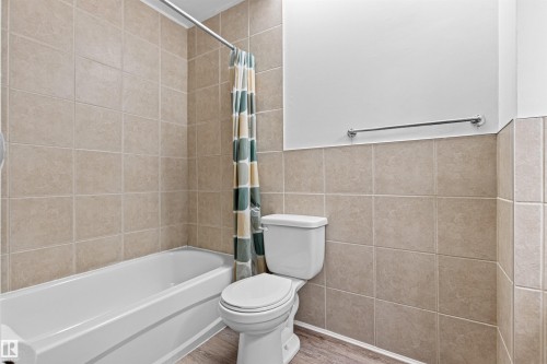 White bathtub with a chrome shower rod, fully tiled walls, and wood-finish flooring - #9 14230 80 St Nw, Edmonton, AB - Indoor Photo Showing Bathroom