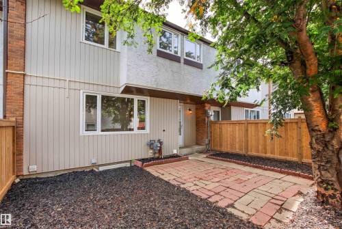 Modern townhome exterior featuring a private patio with brick and gravel hardscaping, wooden privacy fencing, contemporary siding with metal accents, large multi-pane windows, and mature landscaping with established trees providing natural screening - #9 14230 80 St Nw, Edmonton, AB - Outdoor With Exterior