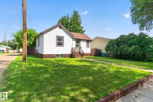 White siding exterior with brown trim, brown shingled roof, front porch with wood stairs, concrete walkway, and a spacious green lawn - 9921 109 Street, Fort Saskatchewan, AB - Outdoor
