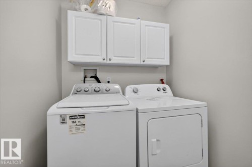 Dedicated laundry room featuring side-by-side washer and dryer with control panels, overhead cabinetry with dual handles for convenient storage, and neutral finishes throughout - 722 88A Street Sw, Edmonton, AB - Indoor Photo Showing Laundry Room