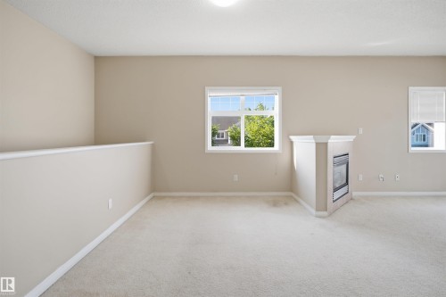 Carpeted upper-level room with a partial wall, featuring a built-in fireplace with a hearth, multiple windows, and light-toned wall finishes - #95 3040 Spence Wd Sw, Edmonton, AB - Indoor Photo Showing Other Room