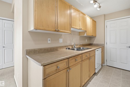Galley-style kitchen featuring wood-finish cabinetry with chrome hardware, laminate countertops, stainless steel appliances including a smooth-top range and sink with single-handle faucet, and neutral ceramic tile flooring - #95 3040 Spence Wd Sw, Edmonton, AB - Indoor Photo Showing Kitchen With Double Sink
