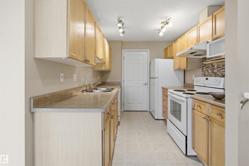 Galley kitchen featuring wood-finish cabinetry, laminate countertops, and a full suite of white appliances - #95 3040 Spence Wd Sw, Edmonton, AB - Indoor Photo Showing Kitchen With Double Sink