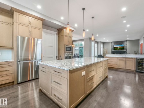 Expansive kitchen island with a light-toned countertop, stainless steel refrigerator, built-in wine cooler, wood-finish flooring, and pendant lighting - 8927 120 Street Nw, Edmonton, AB - Indoor Photo Showing Kitchen With Upgraded Kitchen