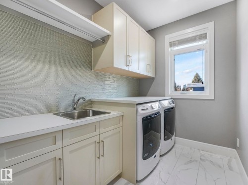 Modern laundry room featuring cream-colored cabinetry with chrome hardware, white quartz countertops, a subway tile backsplash, front-load washer and dryer appliances, and a large window with natural light and outdoor views - 8927 120 Street Nw, Edmonton, AB - Indoor Photo Showing Laundry Room