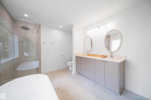 Dual vanity with light-toned cabinetry and oval mirrors, walk-in shower with glass enclosure and rainfall showerhead, and light-toned flooring - #A 12 Invermere Pl, St. Albert, AB - Indoor Photo Showing Bathroom