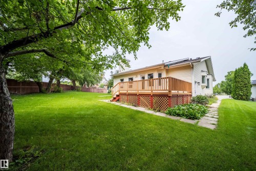 Expansive grassy yard with mature trees, a wood deck featuring lattice skirting, and a bay window on the home's exterior - #A 12 Invermere Pl, St. Albert, AB - Outdoor With Deck Patio Veranda