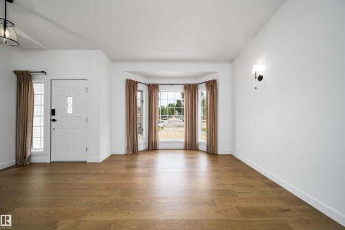 Wood-finish flooring throughout, bay window with multiple panes, white paneled entry door with decorative glass insert, and wall-mounted sconce - #A 12 Invermere Pl, St. Albert, AB - Indoor Photo Showing Other Room