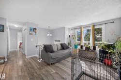Bright living area showcasing wood-finish flooring, large multi-pane windows with natural light, gray upholstered seating, pendant lighting, and access to outdoor space with mature landscaping visible beyond - 