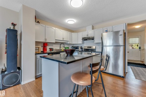 Kitchen featuring an island with a dark countertop, white cabinetry, stainless steel appliances, a white tile backsplash, and wood-finish flooring - 3032 31 Avenue Nw, Edmonton, AB - Indoor Photo Showing Kitchen With Double Sink