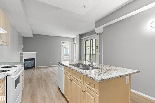 Kitchen island with double basin sink, light wood-finish cabinetry, light-toned stone countertops, and a full-size dishwasher - #404 11103 84 Av Nw, Edmonton, AB - Indoor Photo Showing Kitchen With Double Sink
