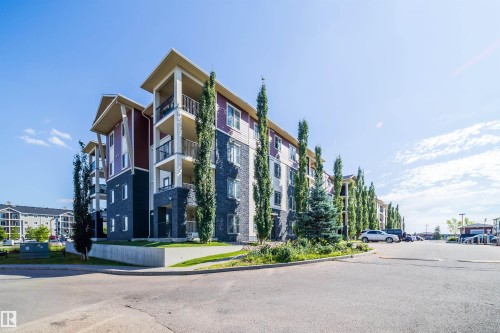 Multi-story building exterior featuring stone and siding, private balconies, abundant mature landscaping, and an asphalt parking area - #315 5816 Mullen Pl Nw, Edmonton, AB - Outdoor
