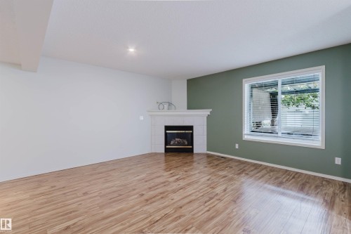 Spacious interior with wood-finish flooring, a corner fireplace featuring a white mantel, and a window with blinds - 1694 Tompkins Wynd Nw, Edmonton, AB - Indoor Photo Showing Other Room With Fireplace