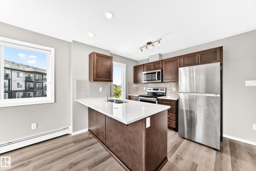 Kitchen featuring wood-finish flooring, dark wood cabinetry, stainless steel appliances, a white island countertop with an integrated sink, and recessed ceiling lighting - #322 148 Ebbers Bv Nw, Edmonton, AB - Indoor Photo Showing Kitchen With Double Sink