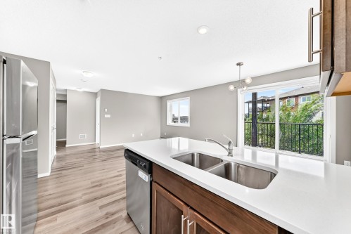 Kitchen island with an undermount double-basin sink, stainless steel appliances, white countertops, and wood-finish cabinetry - #322 148 Ebbers Bv Nw, Edmonton, AB - Indoor Photo Showing Kitchen With Double Sink