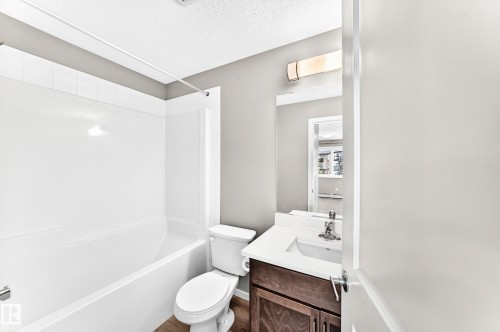Wood-finish flooring, white bathtub with tiled surround, white toilet, vanity with integrated sink and dark wood-finish cabinetry, and wall-mounted light fixture - #322 148 Ebbers Bv Nw, Edmonton, AB - Indoor Photo Showing Bathroom