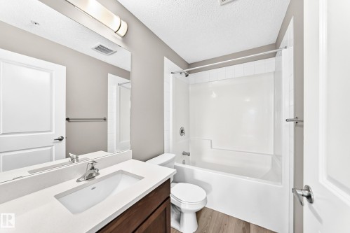 Wood-finish flooring, single vanity with a white countertop and integrated sink, chrome faucet, wall-mounted mirror, and a white bathtub with a shower surround - #322 148 Ebbers Bv Nw, Edmonton, AB - Indoor Photo Showing Bathroom