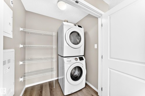 Laundry area featuring a stacked washer and dryer, wire shelving, wood-finish flooring, and a flush mount ceiling light - #322 148 Ebbers Bv Nw, Edmonton, AB - Indoor Photo Showing Laundry Room