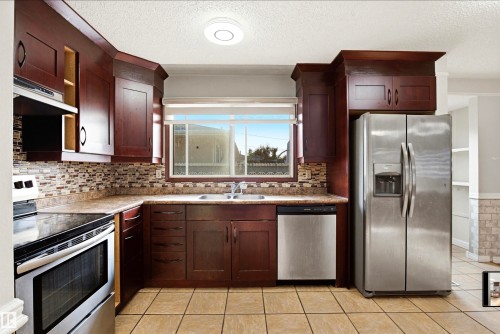 Wood-finish cabinetry paired with stainless steel appliances, a mosaic tile backsplash, and a ceramic tile floor - 13604 90A Street Nw, Edmonton, AB - Indoor Photo Showing Kitchen With Stainless Steel Kitchen With Double Sink