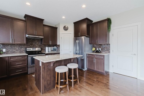 Kitchen featuring rich wood cabinetry, stainless steel appliances, stone countertops, a tiled backsplash, and wood-finish flooring - 3843 Agar Green Sw, Edmonton, AB - Indoor Photo Showing Kitchen With Stainless Steel Kitchen With Upgraded Kitchen