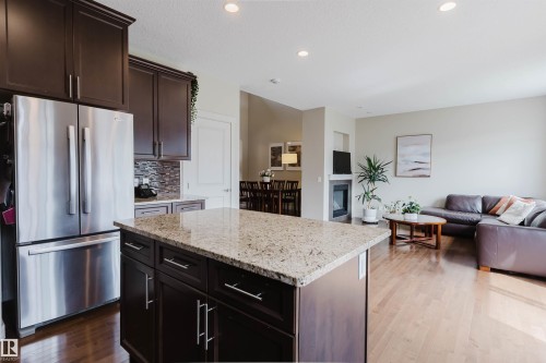Dark wood cabinetry with brushed nickel hardware, stainless steel refrigerator, granite-style kitchen island, wood-finish flooring, and a built-in fireplace - 3843 Agar Green Sw, Edmonton, AB - Indoor Photo Showing Kitchen With Stainless Steel Kitchen