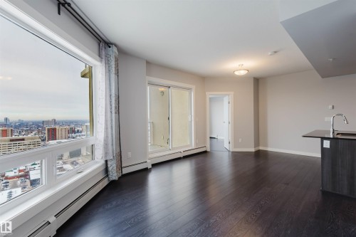 Expansive window with city scenery, dark wood-finish flooring, sliding glass door to exterior, integrated kitchen island with sink, and recessed ceiling lighting - #2207 10226 104 St Nw, Edmonton, AB - Indoor