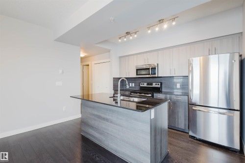 Kitchen featuring an island with a dark countertop and undermount sink, stainless steel appliances, light wood-finish cabinetry, and dark wood-finish flooring - #2207 10226 104 St Nw, Edmonton, AB - Indoor Photo Showing Kitchen With Stainless Steel Kitchen