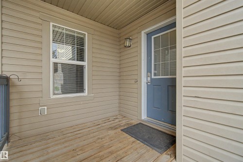 Covered front porch with wood-finish decking, tan siding, decorative wall sconce, and a blue entry door with a divided light window - 1602 Chapman Way Sw, Edmonton, AB - Outdoor With Exterior