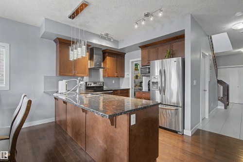 Modern kitchen with wood-finish flooring, stainless steel appliances, dark wood cabinetry, a central island with a stone countertop, and pendant lighting - 1602 Chapman Way Sw, Edmonton, AB - Indoor Photo Showing Kitchen With Double Sink