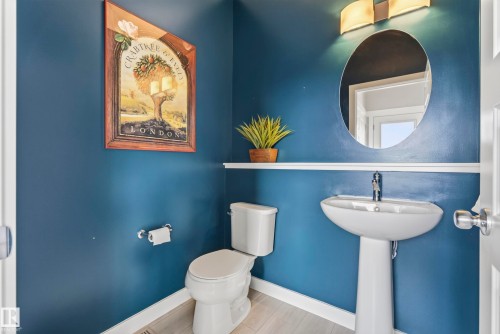 Powder room featuring a pedestal sink with chrome faucet, white ceramic toilet, and rich blue walls accented by a round mirror with brushed metal frame - 45 Joyal Way, St. Albert, AB - Indoor Photo Showing Bathroom