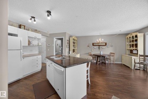 Open-concept kitchen featuring white cabinetry, a dark countertop island with a double sink, and wood-finish flooring - #7 155 Crocus Cr, Sherwood Park, AB - Indoor Photo Showing Kitchen With Double Sink
