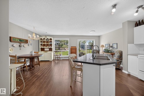 Open-concept living area featuring wood-finish flooring, a kitchen island with a double sink and dark countertop, and white kitchen cabinetry - #7 155 Crocus Cr, Sherwood Park, AB - Indoor Photo Showing Kitchen With Double Sink