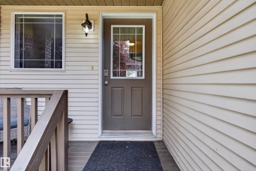 Front entryway featuring a brown door with decorative glass, beige horizontal siding, a white-framed window, an exterior wall sconce, and a wood-finish railing - #7 155 Crocus Cr, Sherwood Park, AB - Outdoor With Exterior