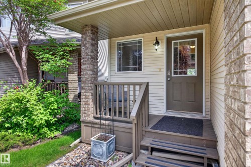 Front entrance featuring a dark brown door with decorative glass, beige siding, a brick column, a wood-toned porch with railing, and integrated exterior lighting - #7 155 Crocus Cr, Sherwood Park, AB - Outdoor With Deck Patio Veranda With Exterior