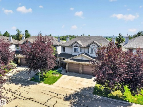 Two-story residence featuring a light-colored exterior, an attached two-car garage with wood-finish doors, a pitched roofline, and front landscaping with mature trees - #7 155 Crocus Cr, Sherwood Park, AB - Outdoor With Facade