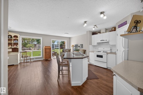 Open-concept kitchen and living area featuring wood-finish flooring, white cabinetry, a kitchen island with a double sink, and a sliding glass patio door to the backyard - #7 155 Crocus Cr, Sherwood Park, AB - Indoor Photo Showing Kitchen With Double Sink